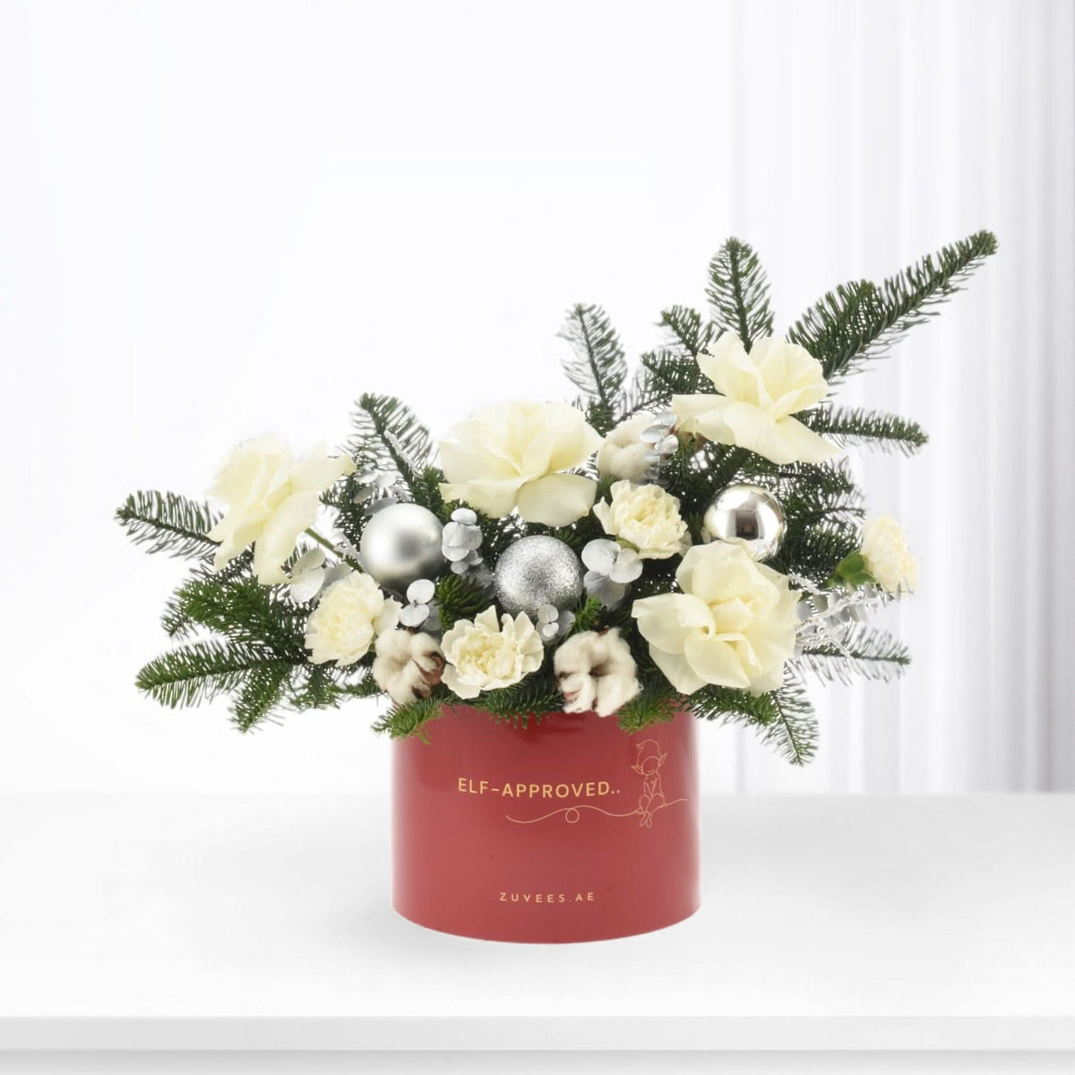 Red holiday floral arrangement with white blooms, silver ornaments, and pine in festive container.