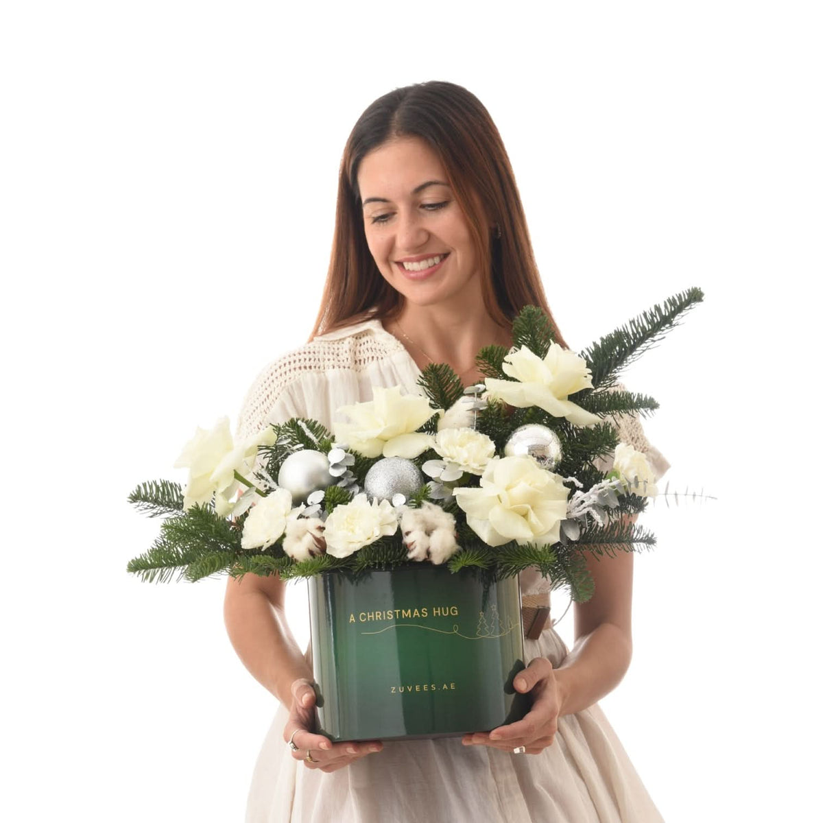 Woman smiles holding festive green box with white flowers, pine, and silver ornaments.