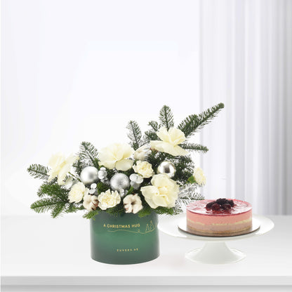Elegant green holiday bouquet with white blooms, pine, and silver ornaments beside layered berry cake.
