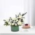 Elegant green holiday bouquet with white blooms, pine, and silver ornaments beside layered berry cake.