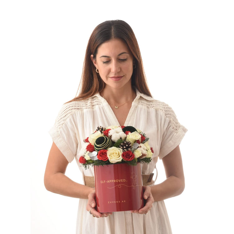 Woman holds festive red box bouquet with red/white roses, pinecones, and "Elf-Approved" text.