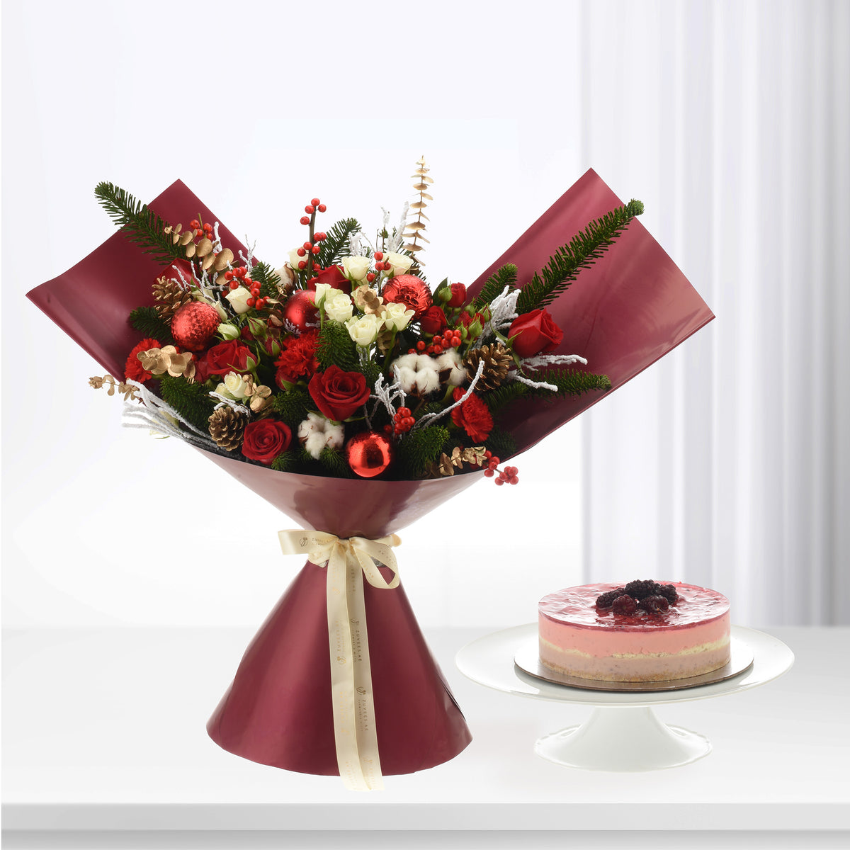 Vibrant red roses, pine, berries, and ornaments in burgundy wrap beside pink berry cake.