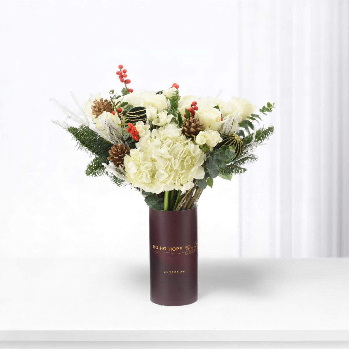 White roses and hydrangeas in burgundy tube with festive red berries and pinecones.