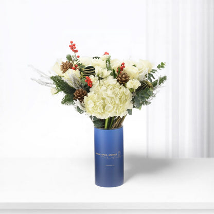 Elegant white floral arrangement in blue cylinder vase with festive pine and berries.