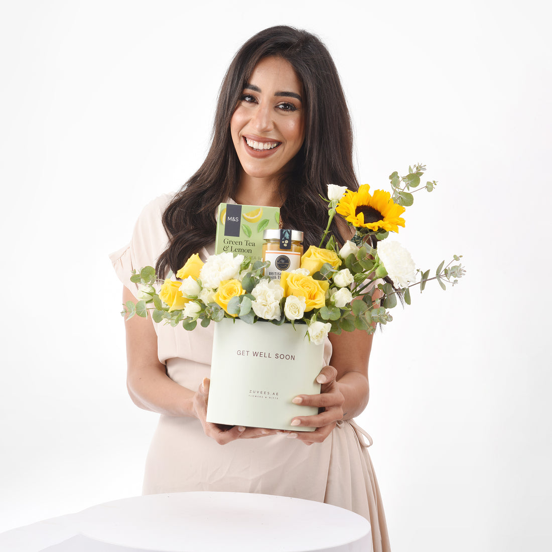 Smiling woman holds “Get Well Soon” gift box with yellow/white roses, sunflower, tea, honey.