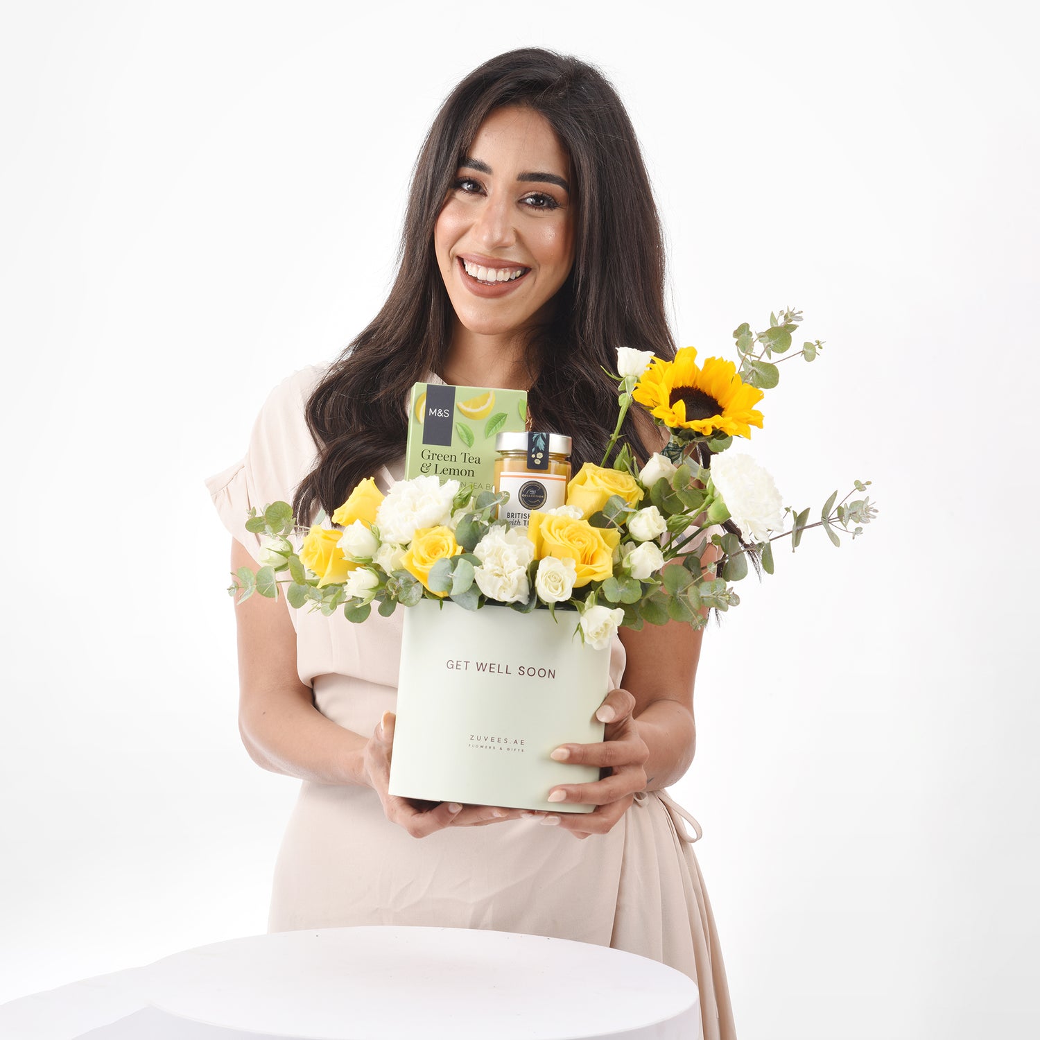 Smiling woman holds “Get Well Soon” gift box with yellow/white roses, sunflower, tea, honey.