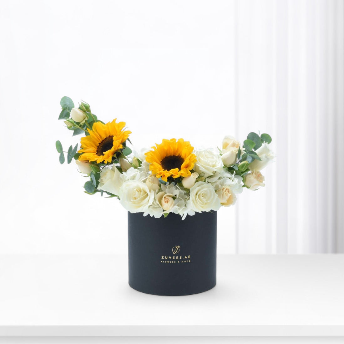 Vibrant sunflowers and white roses in sleek black box with gold logo.