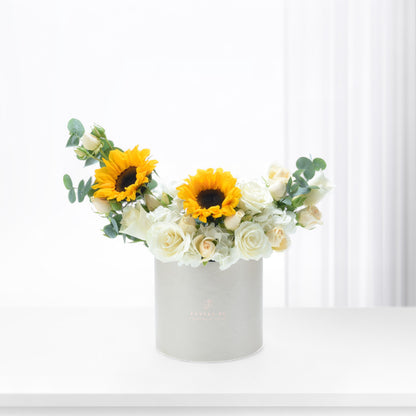 White cylindrical box holds vibrant sunflowers and creamy roses against bright background.