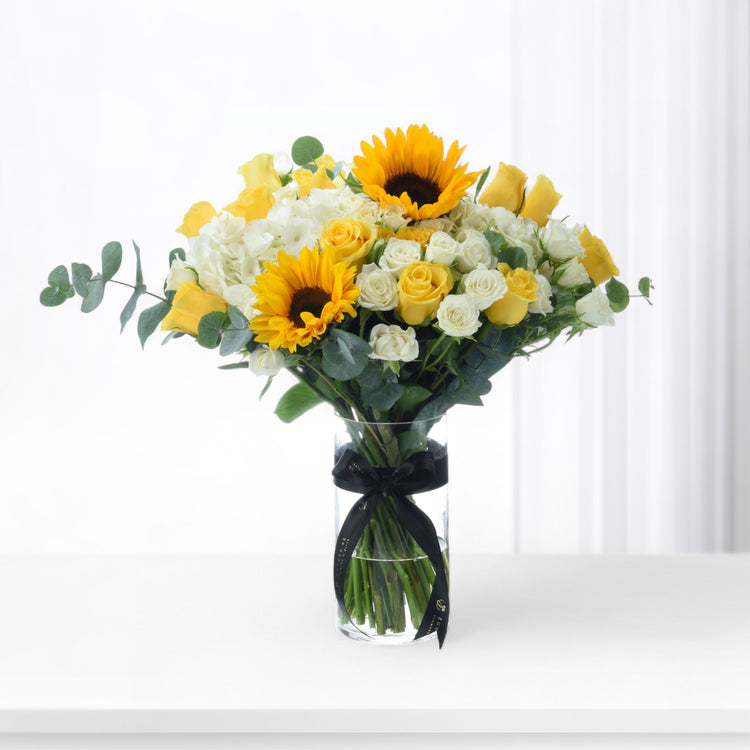 Vibrant yellow sunflowers and roses in clear vase with black ribbon.