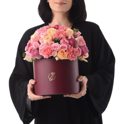 Elegant burgundy hat box filled with lush pink and peach roses, held by a smiling woman.