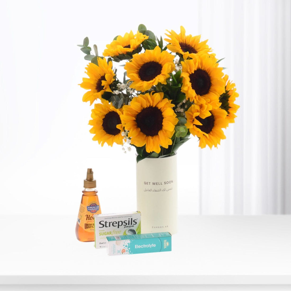 Vibrant sunflower bouquet in white vase with "Get Well Soon" text, beside honey, Strepsils, and electrolyte.