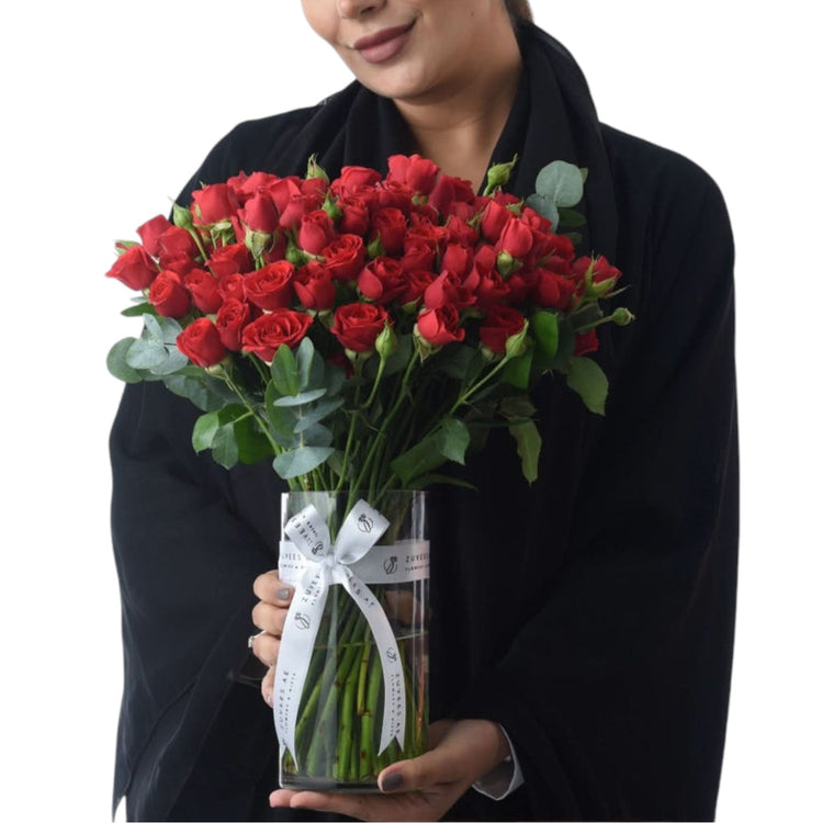 Vibrant red roses in clear vase with white ribbon, held by smiling woman in black.