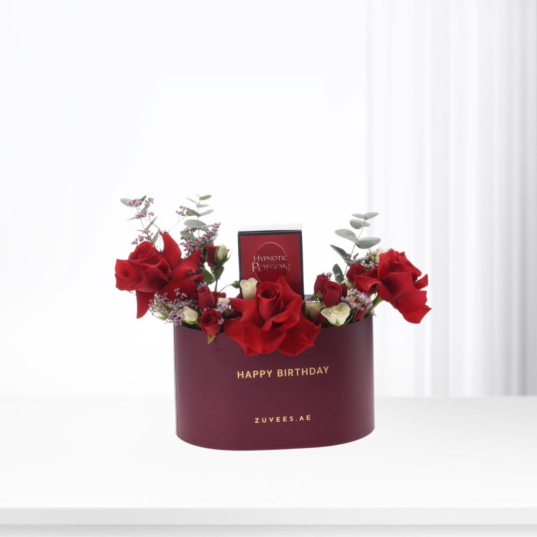 Deep burgundy box with gold "Happy Birthday" text, overflowing with vibrant red roses and delicate white blooms, accented by greenery and a "Hypnotic Poison" perfume bottle.