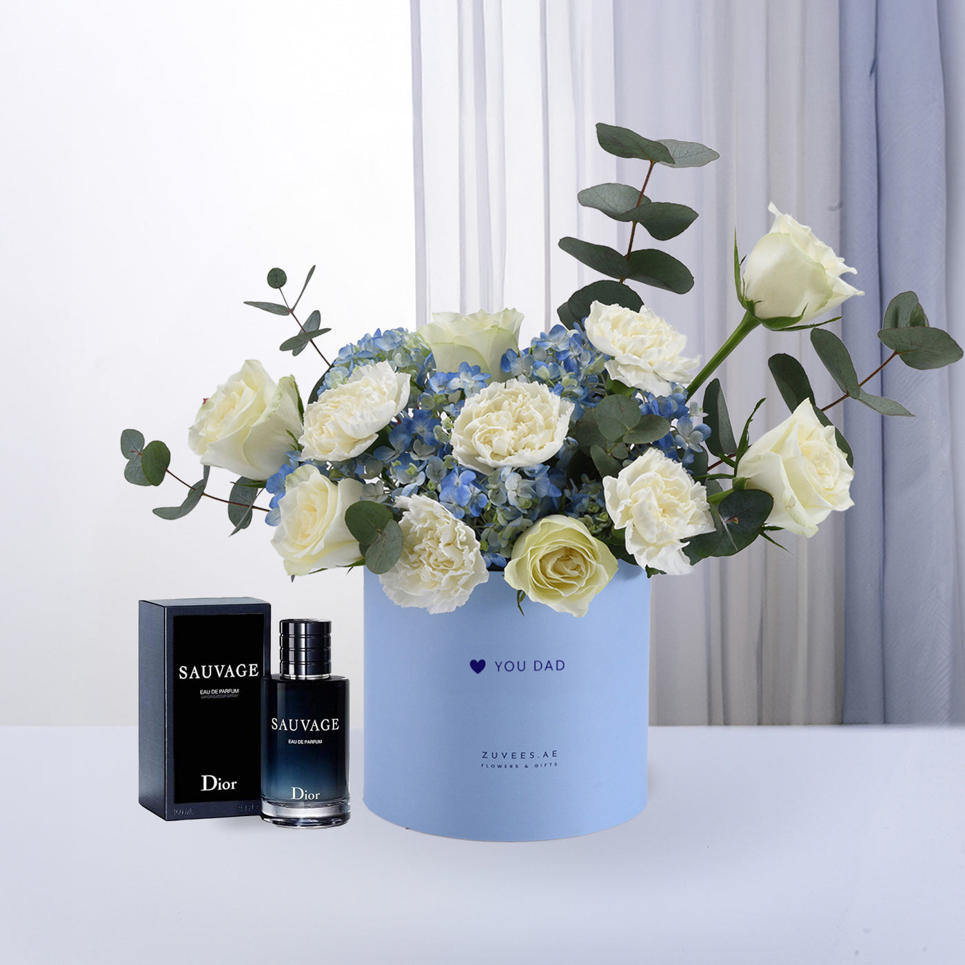 Blue box holds white roses and blue hydrangeas, beside Dior Sauvage perfume.