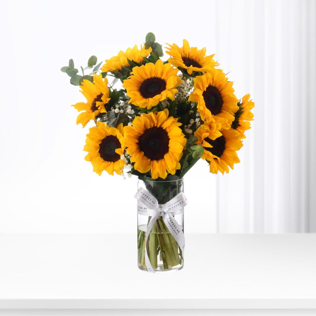 Vibrant sunflowers in clear vase with white ribbon against bright white background.