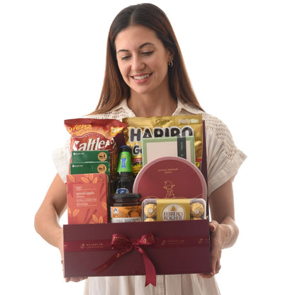 Woman smiles holding rich burgundy gift box filled with assorted treats and chocolates.