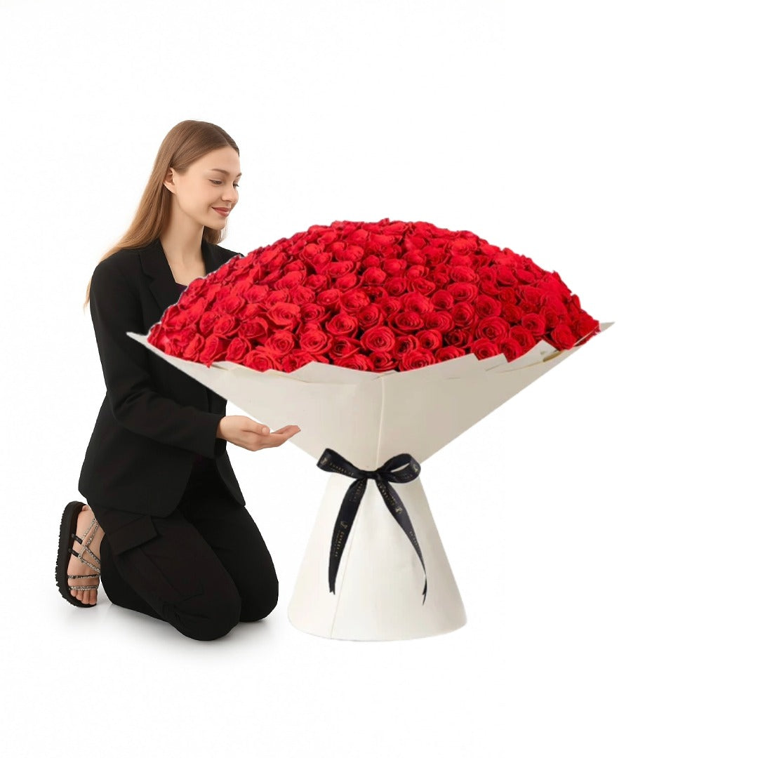 Woman kneels beside massive red rose bouquet in elegant white wrapping with black ribbon.