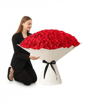 Woman kneels beside massive red rose bouquet in elegant white wrapping with black ribbon.