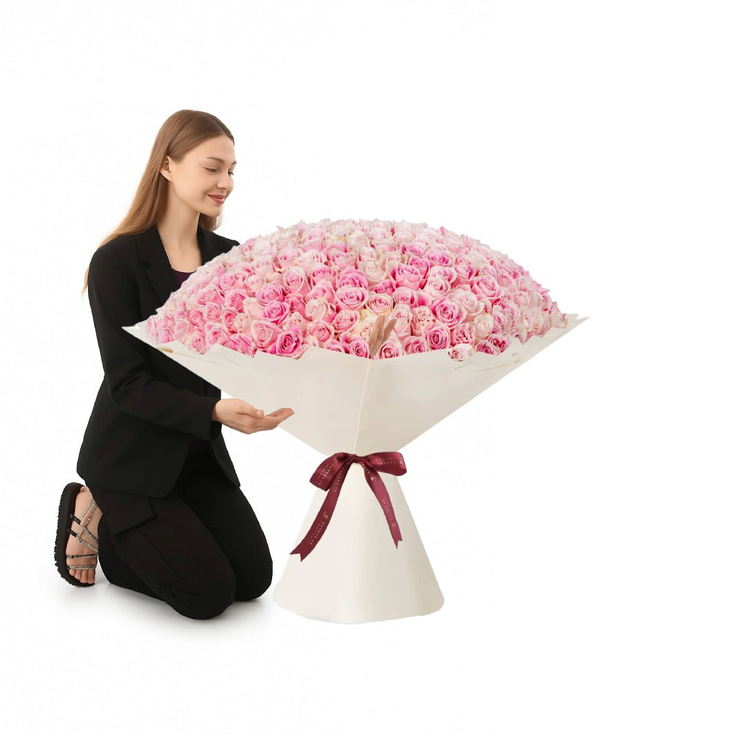 Woman kneels beside massive pink rose bouquet wrapped in white paper with burgundy ribbon.