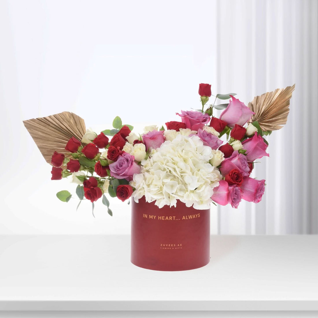 Vibrant red, pink, and white roses with hydrangeas in a deep red cylindrical vase.