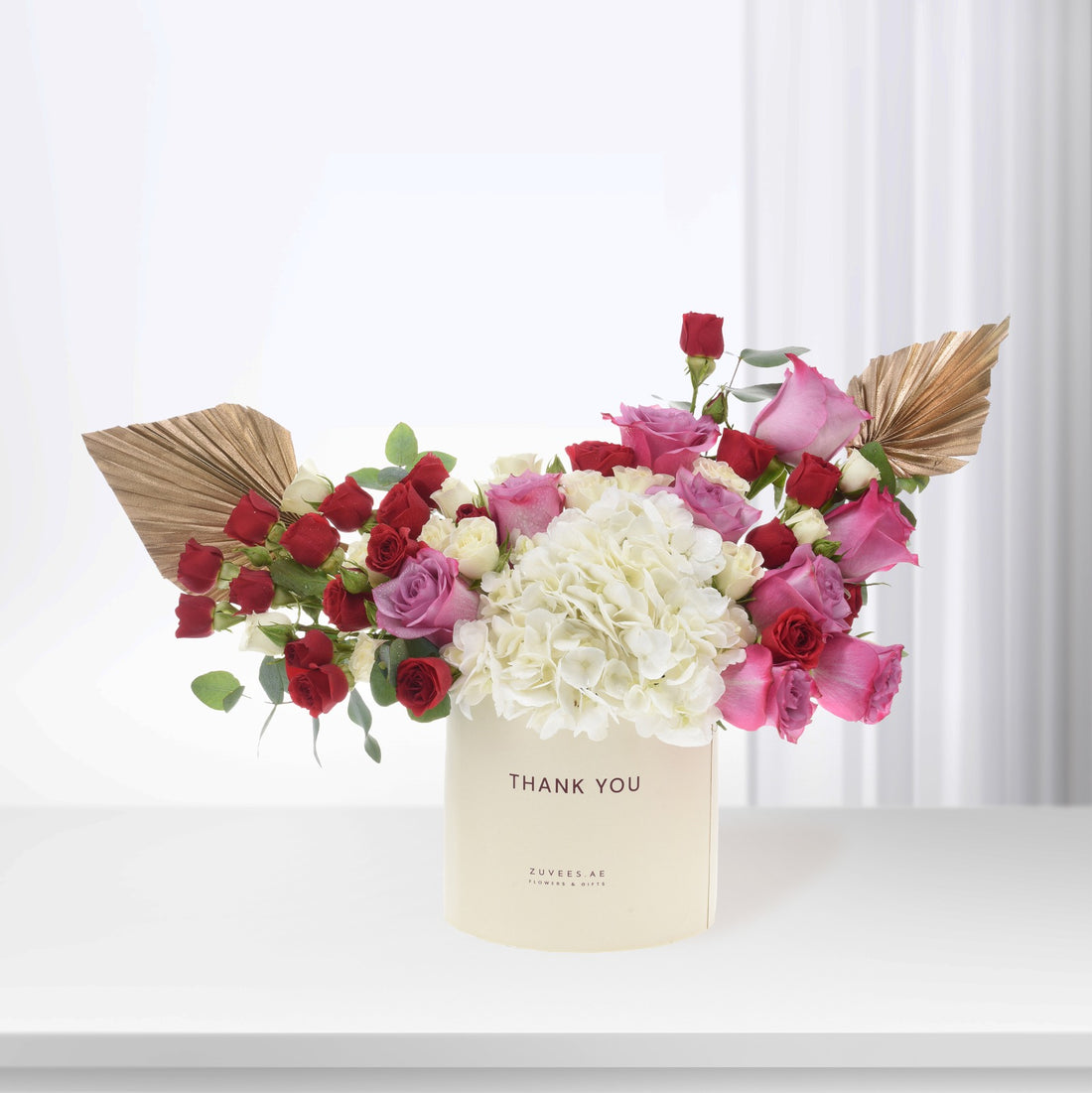 Cream box holds vibrant red, pink, white roses and hydrangeas, accented with gold fan leaves.