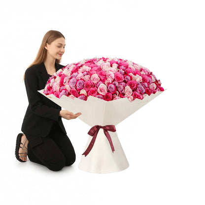 Woman kneels beside massive, vibrant pink rose bouquet in elegant white wrap with burgundy bow.