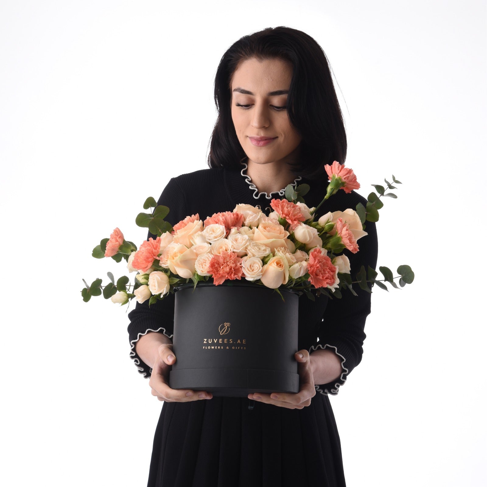 Elegant woman holds black box with peach roses and carnations, eucalyptus, branded ZUVEES.AE.