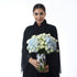 Elegant woman in black holds vase of white roses and blue hydrangeas, tied with white ribbon.