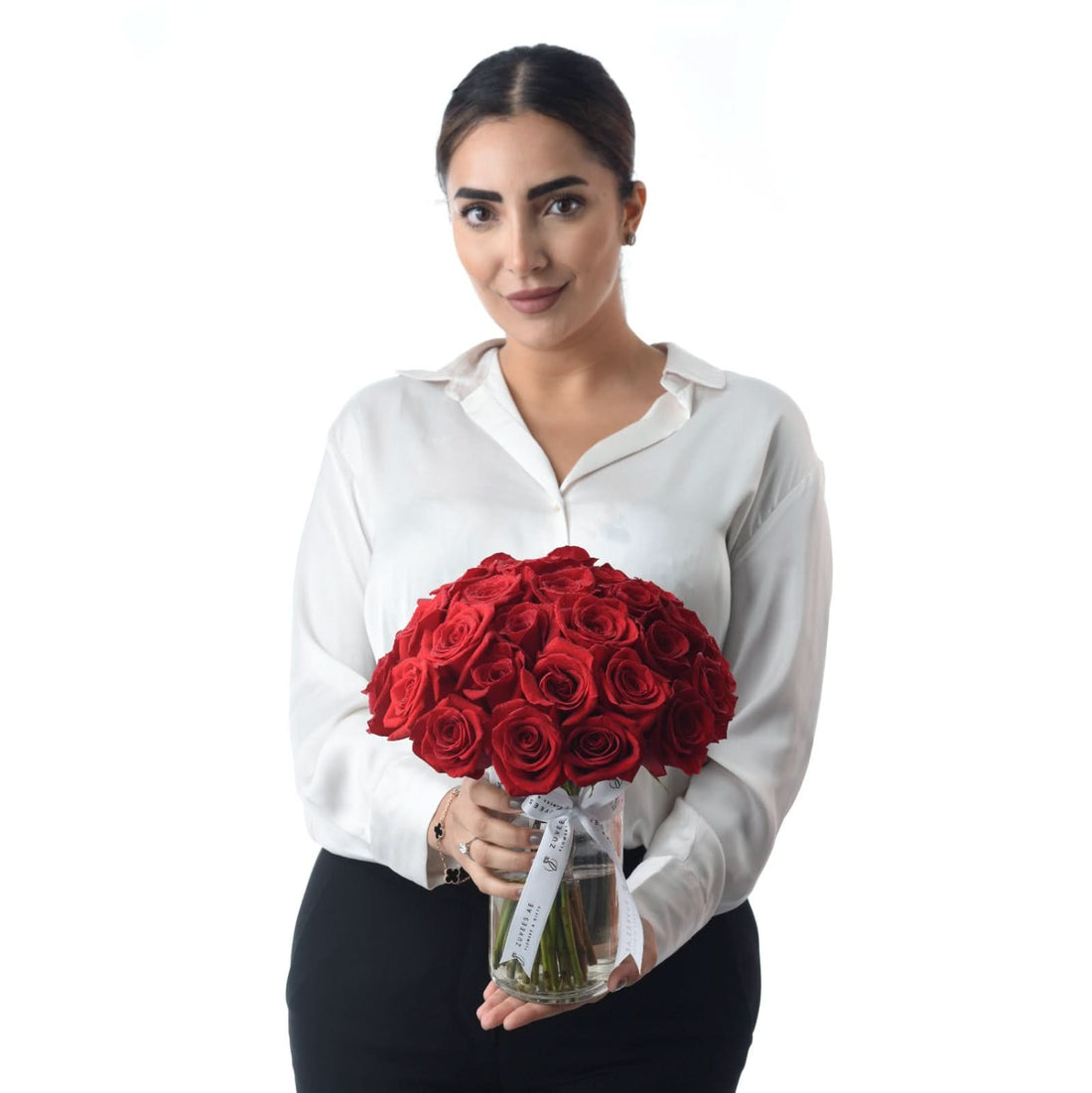Woman in white blouse holds vibrant red roses in clear glass vase.