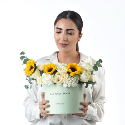 Woman holds mint box with sunflowers and white roses saying “Get Well Soon.”