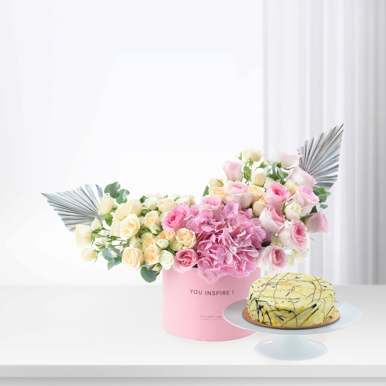 Pink floral arrangement with silver accents beside yellow cake on white stand.
