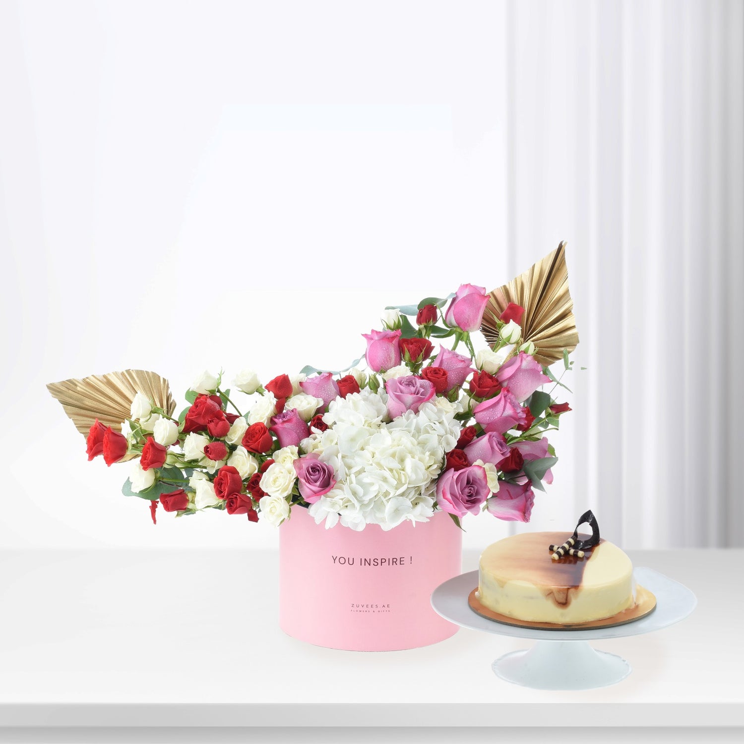 Pink box holds vibrant red, pink, white roses and hydrangeas, accented with gold fans, beside a caramel-glazed cake.