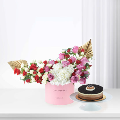 Pink box holds vibrant red, pink, white roses and hydrangeas, accented with gold fans. Beside, layered chocolate cake on white stand.