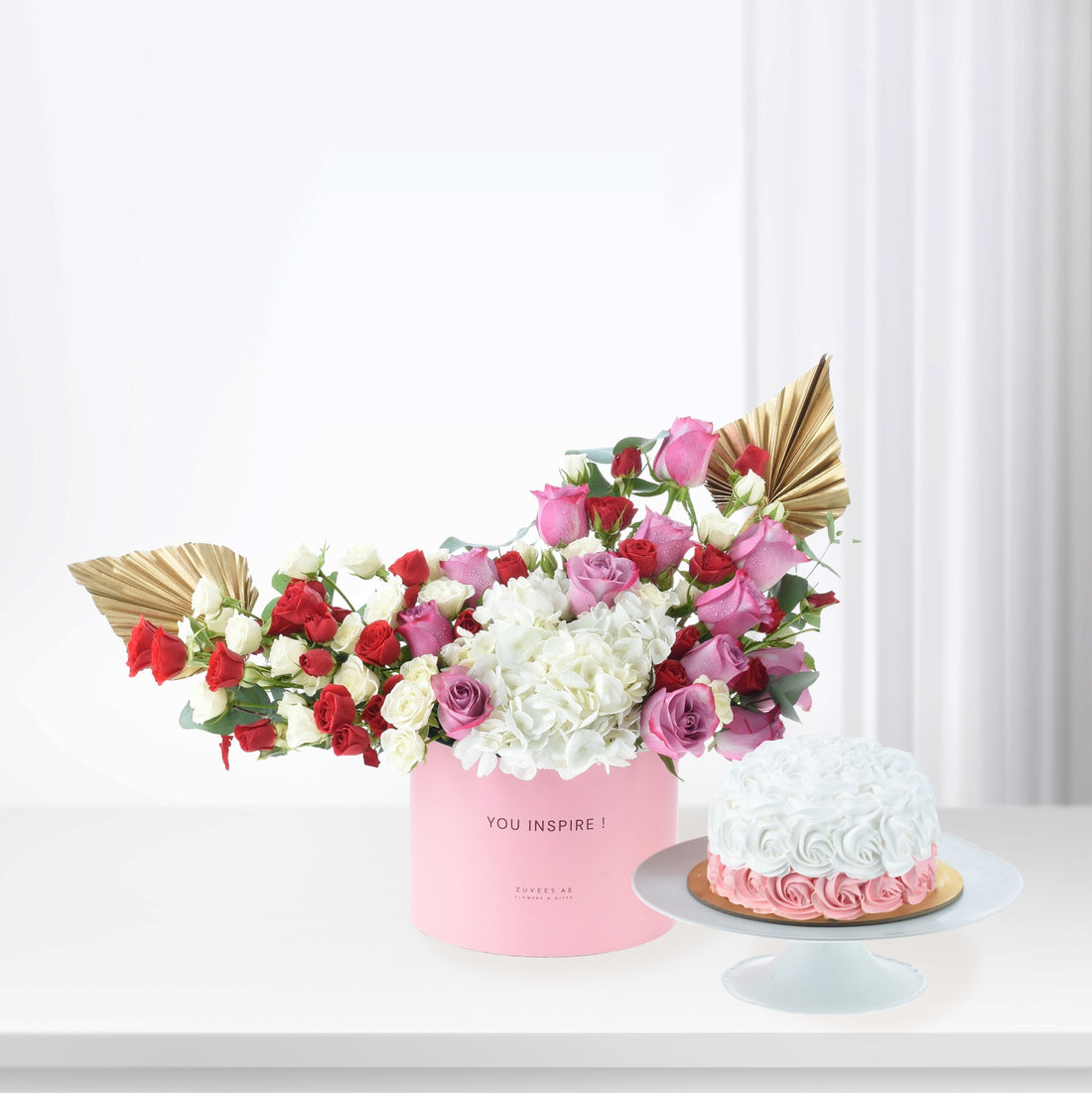 Pink box holds red, pink, white roses and hydrangeas, beside cake with rose icing.