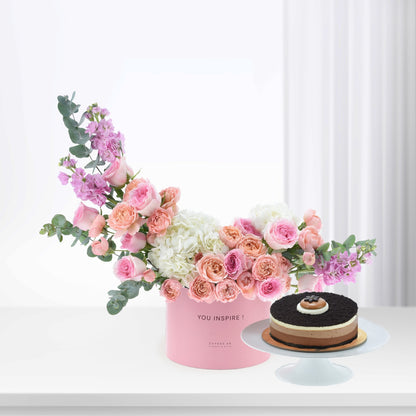 Pink floral arrangement with roses, hydrangeas, eucalyptus in "YOU INSPIRE!" box beside chocolate cake.