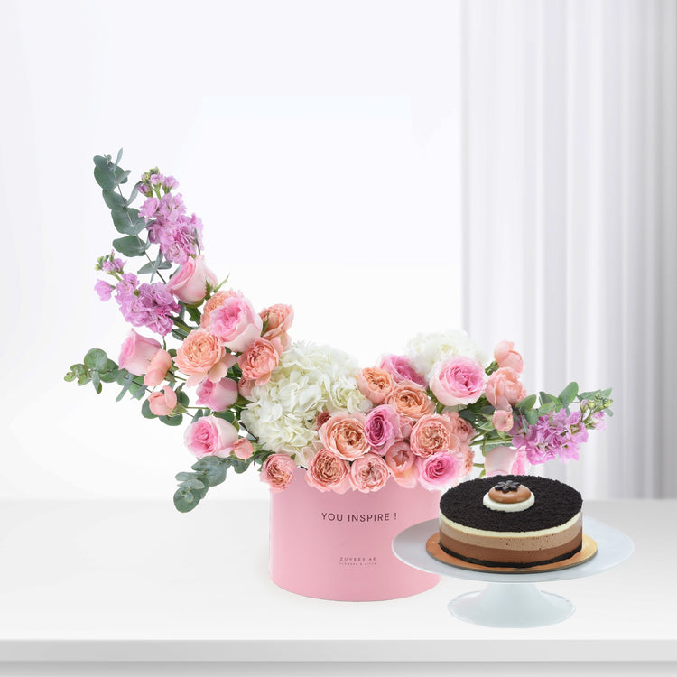 Pink floral arrangement with roses, hydrangeas, eucalyptus in "YOU INSPIRE!" box beside chocolate cake.