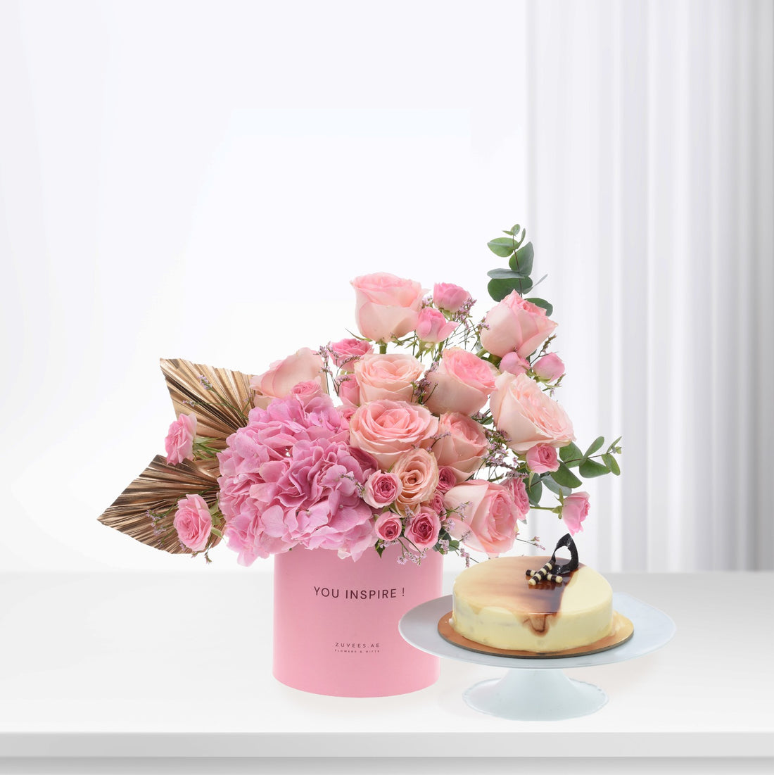 Pink roses, hydrangeas in "YOU INSPIRE!" box beside caramel cheesecake, gold accents, soft white background.