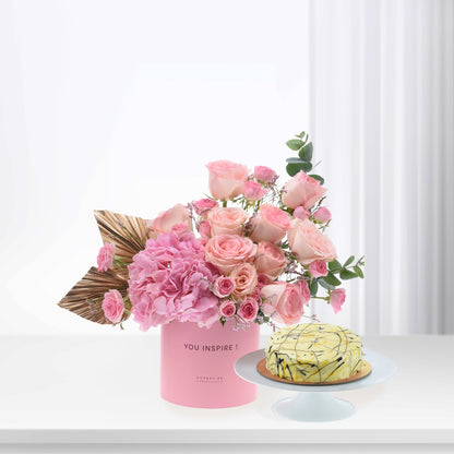 Pink roses, hydrangeas in "YOU INSPIRE!" box beside yellow cake with chocolate drizzle, gold leaf accents.