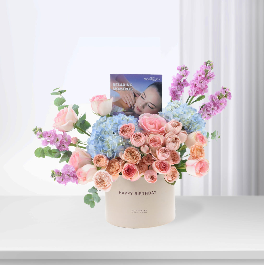 Pastel pink roses, blue hydrangeas, and purple blooms in cream box with "Happy Birthday" text.