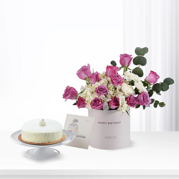 Pink roses and white hydrangeas in a "Happy Birthday" box beside a coconut cake.