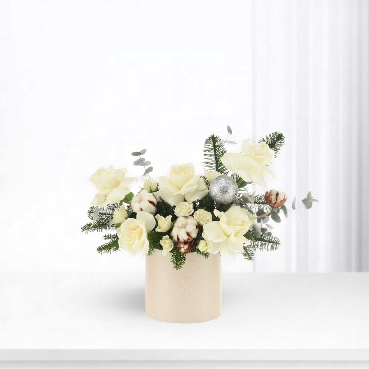 Cream roses, cotton, pine, silver ornament in beige vase; elegant winter floral arrangement.