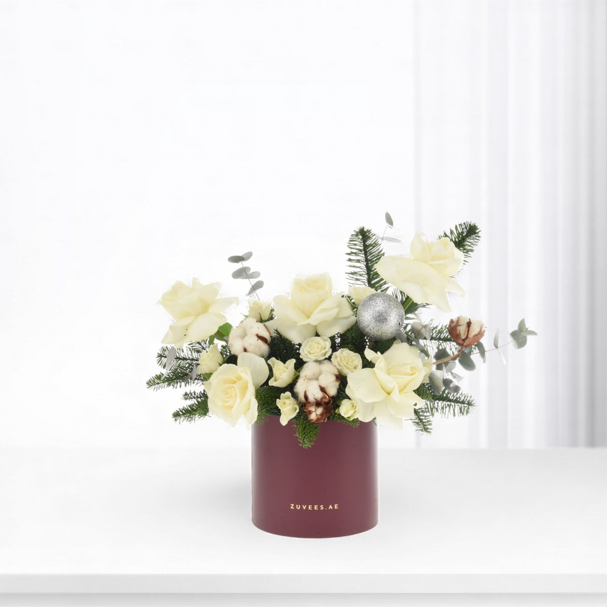 White roses, cotton, and evergreens in burgundy vase with silver ornament.