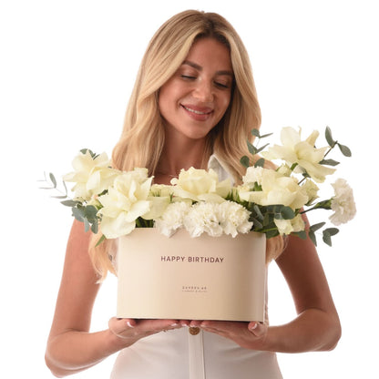 Blonde woman holds elegant cream box with white roses, carnations, eucalyptus, "Happy Birthday" text.
