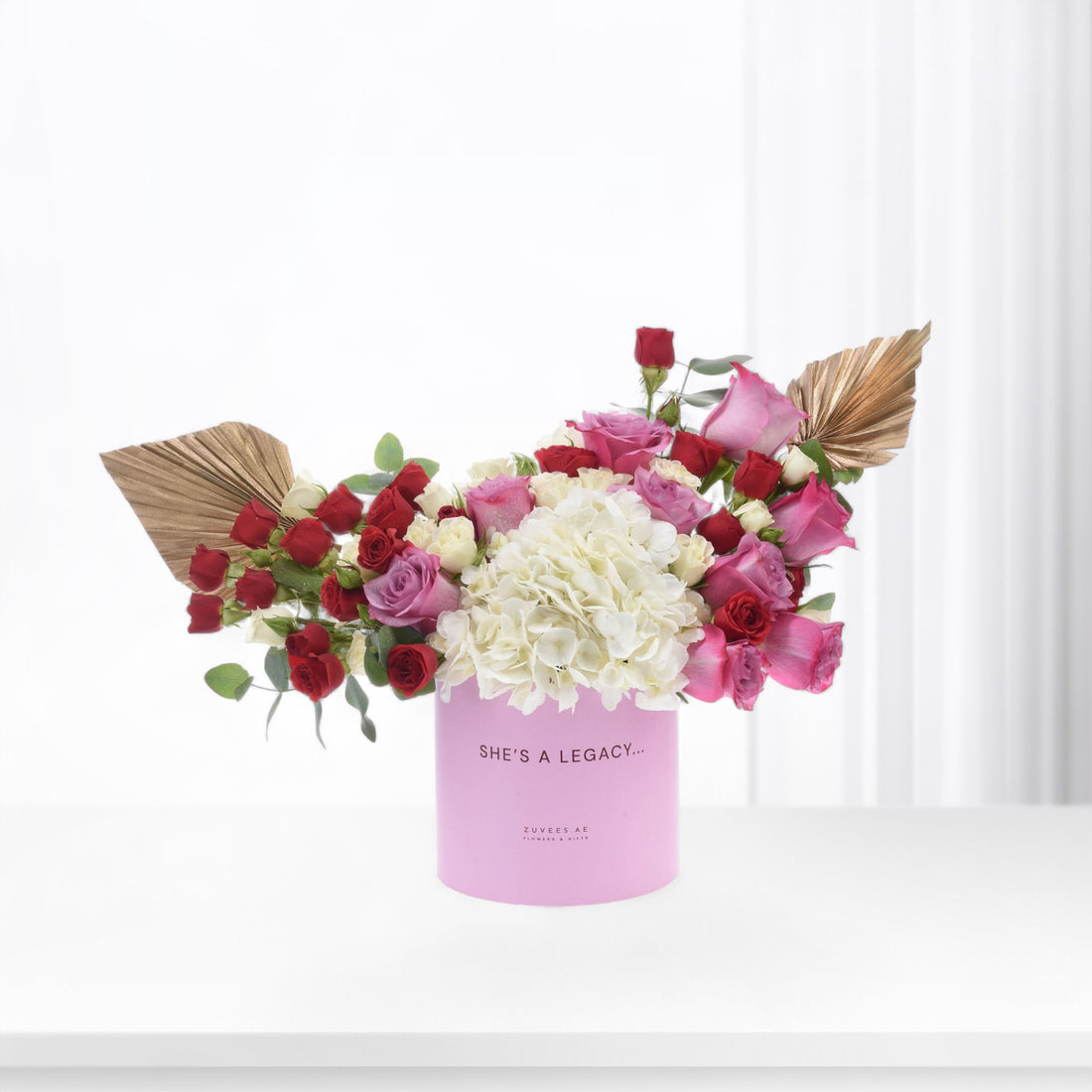 Pink box holds vibrant red, pink, and white roses with gold leaf accents.