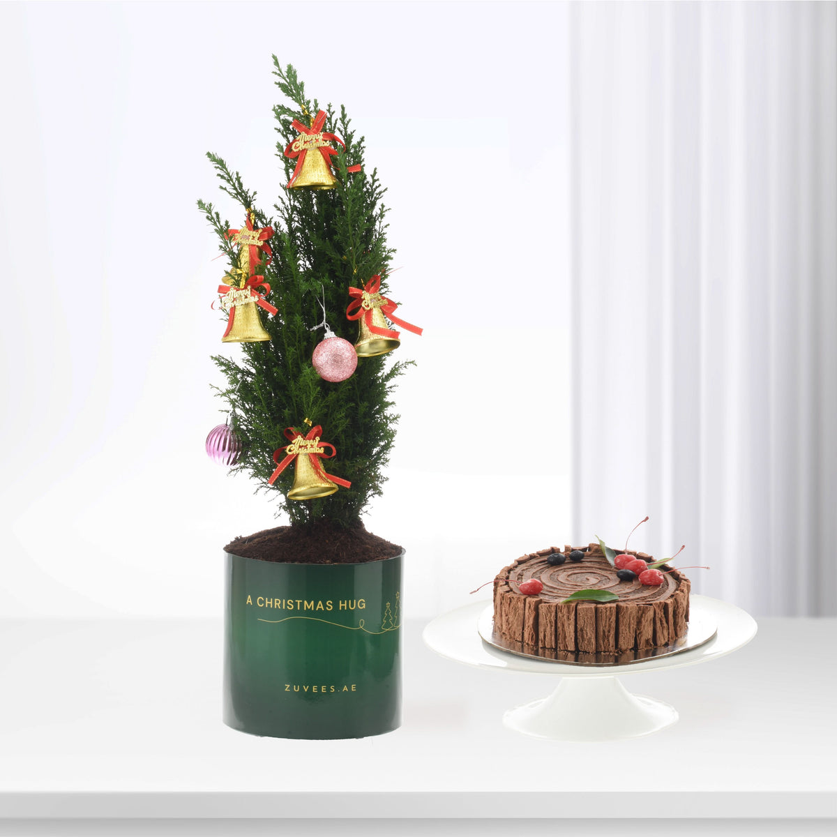 Mini Christmas tree in green pot with gold bells, pink ornaments, beside chocolate Yule log cake.