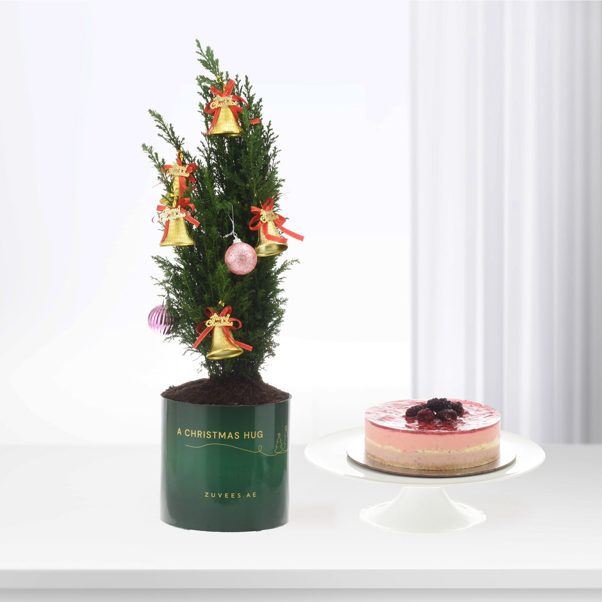 Mini Christmas tree with gold bells and pink baubles beside berry-topped cake.
