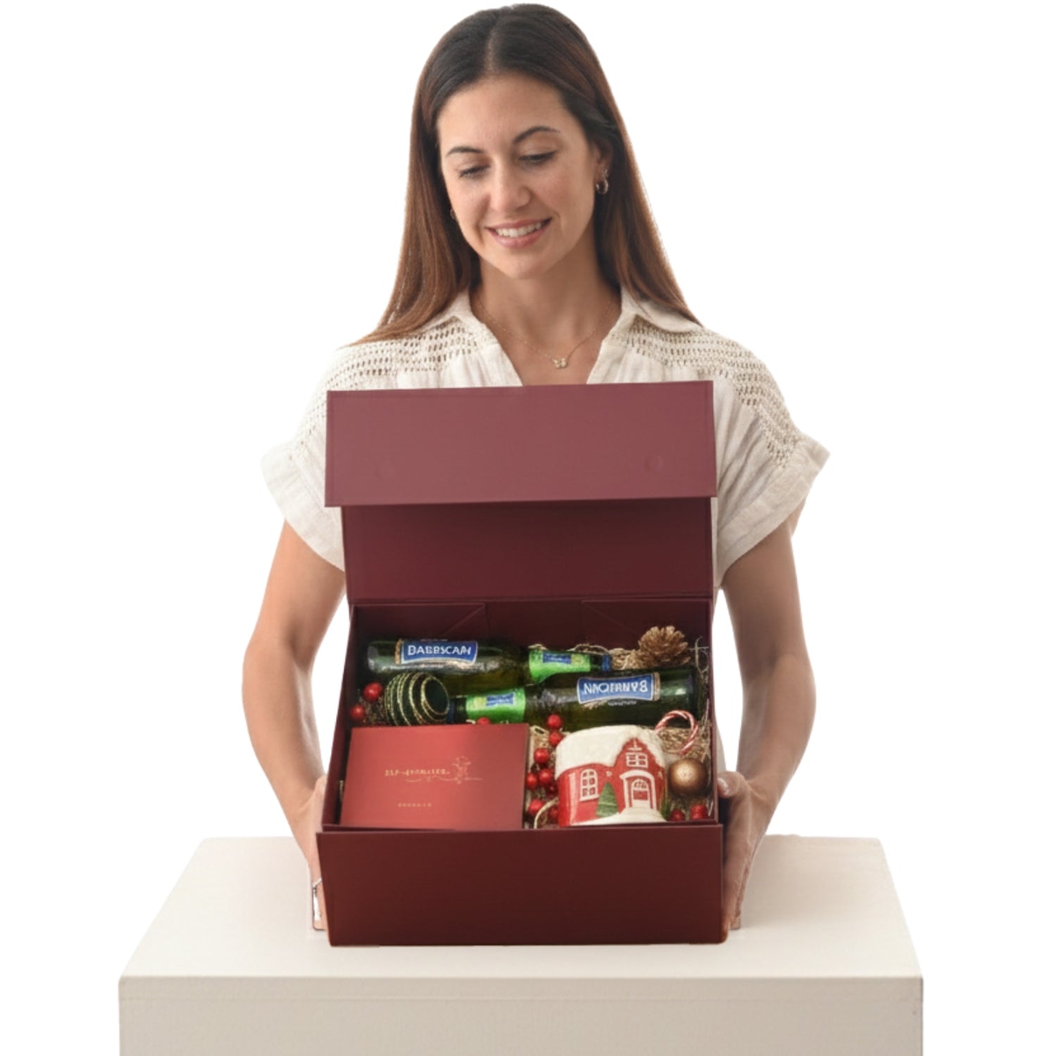 Woman smiles holding open burgundy gift box revealing festive wine, mug, and holiday decor.