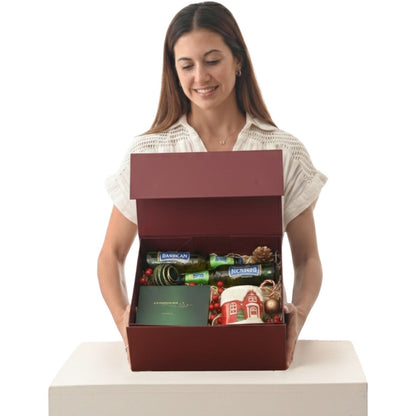 Woman holds festive red gift box with wine, ornaments, and holiday mug inside.