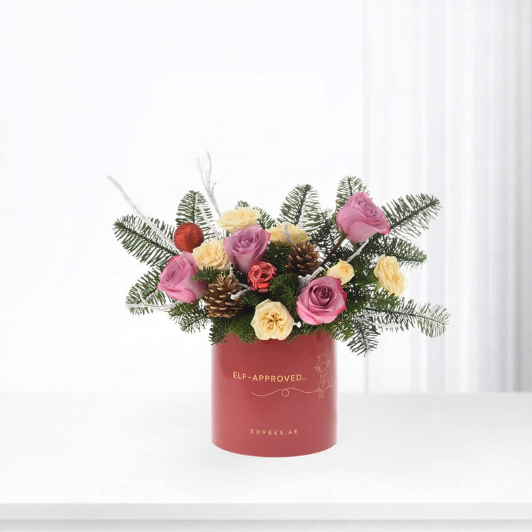 Festive red vase with pink, yellow roses, pine, ornaments, "Elf-Approved" gold text, holiday floral arrangement.