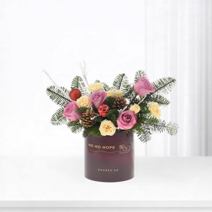 Purple vase holds pink, yellow roses, pine, pinecones, red ornaments, festive "Ho Ho Hope" text.